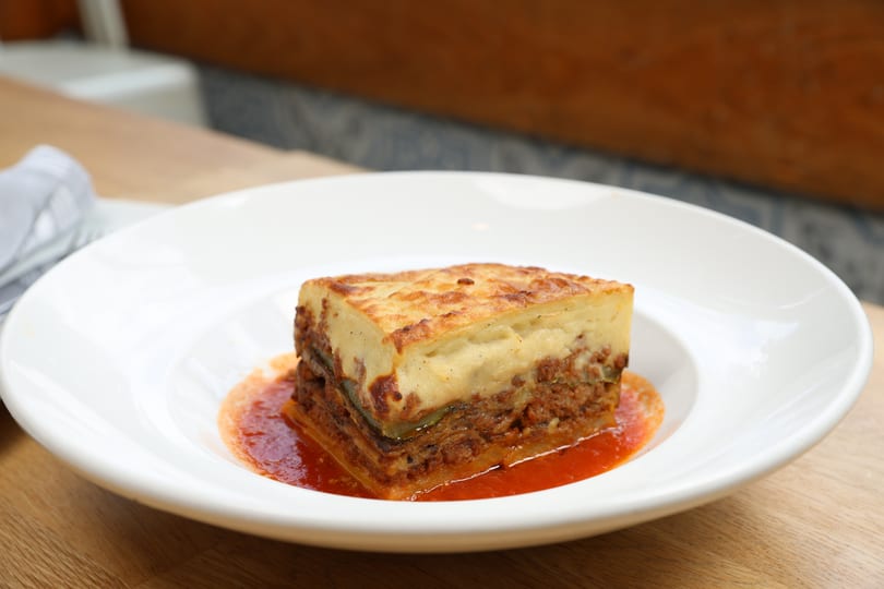 A square slice of moussaka with a golden-brown top, served in a shallow white bowl with red sauce.