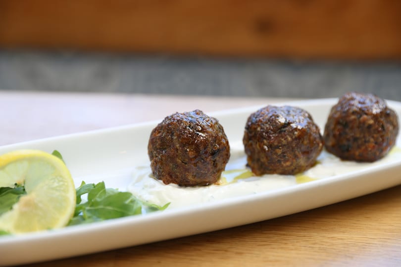 Three meatballs on a white plate with a dollop of sauce, a lemon wedge, and greens.