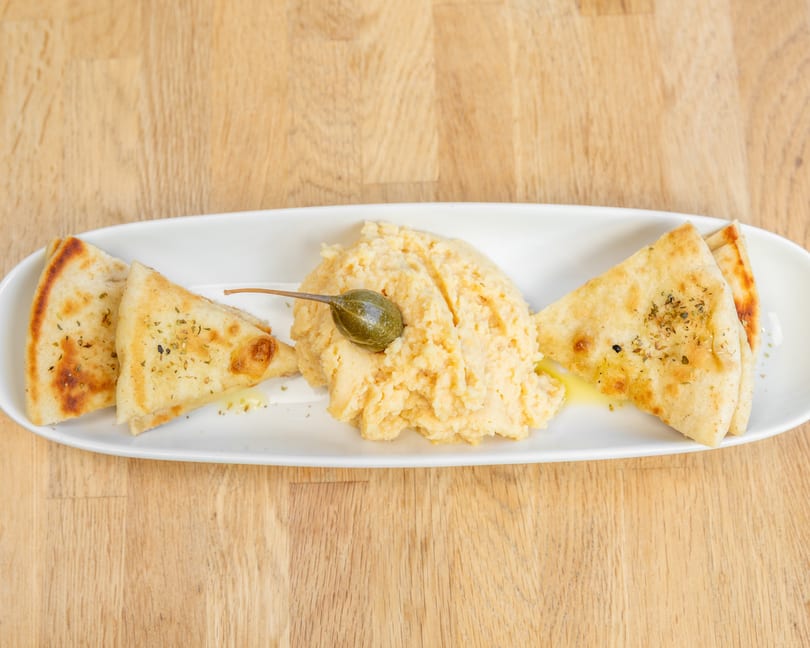 Hummus with a caper on top, served with pita bread triangles drizzled with olive oil.