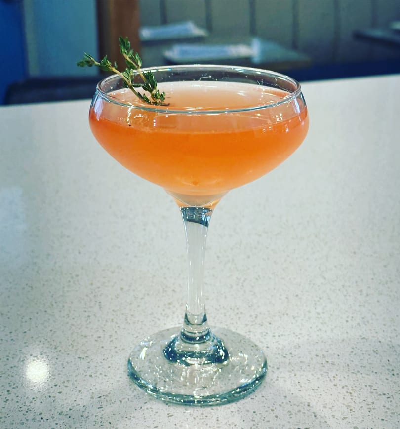 A vibrant orange cocktail in a coupe glass, garnished with a sprig of thyme on a white speckled surface.
