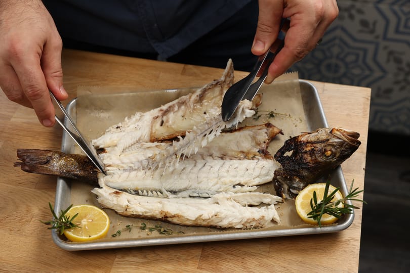 Hands using tongs to debone a whole grilled fish on a baking sheet with lemon and rosemary.