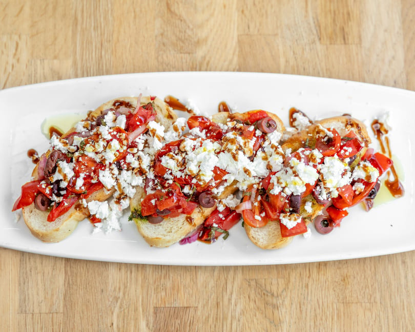 Four bruschetta pieces topped with diced tomatoes, feta, olives, and balsamic glaze on a white plate.