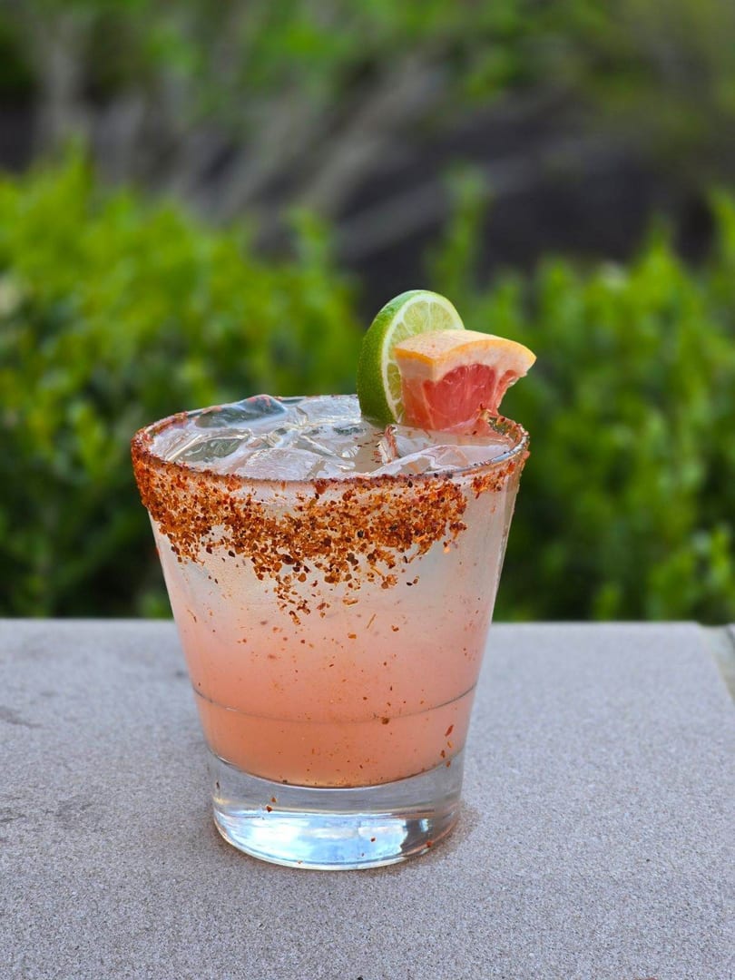 Pink cocktail with ice, a chili-lime rim, and garnished with lime and grapefruit slices.