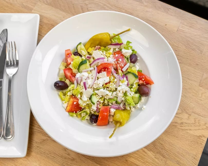 A vibrant Greek salad with lettuce, tomatoes, cucumbers, feta, olives, and pepperoncini on a white plate.