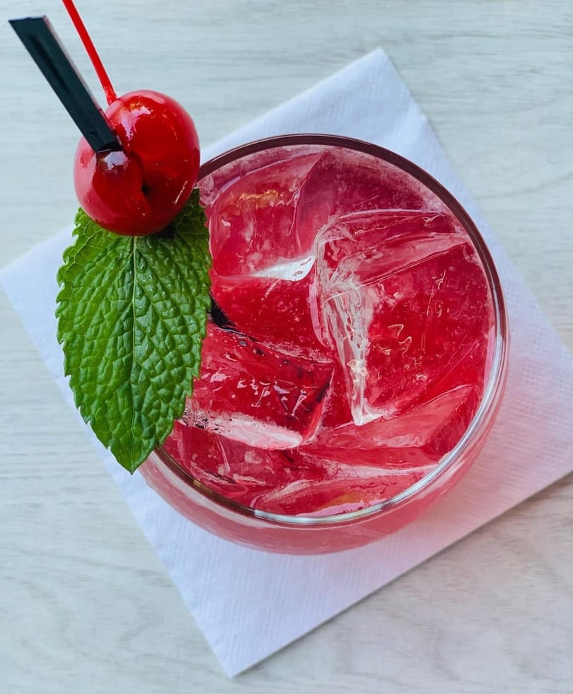 Top-down view of a vibrant red cocktail with ice, garnished with a cherry and mint leaf.