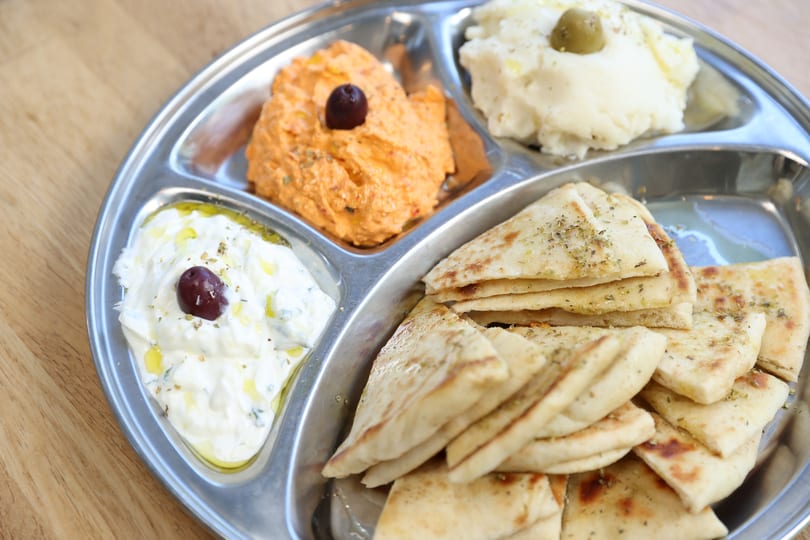 A metal platter with four compartments holding pita bread, tzatziki, hummus, and mashed potatoes.