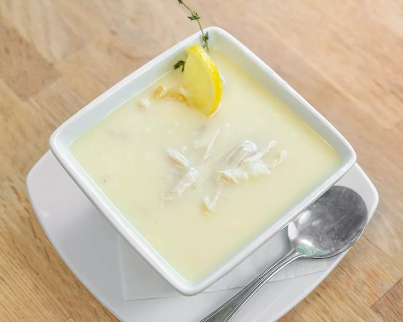 Creamy avgolemono soup with chicken, garnished with a lemon slice and thyme, on a wooden table.